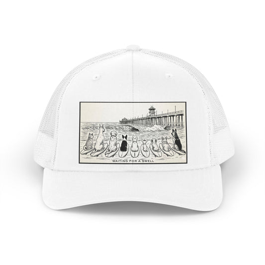 White trucker hat with a picture of a pack of nine dogs sitting on their surfboards, facing the ocean, with the Huntington Beach pier in the background. It says Waiting for a Swell at the bottom.