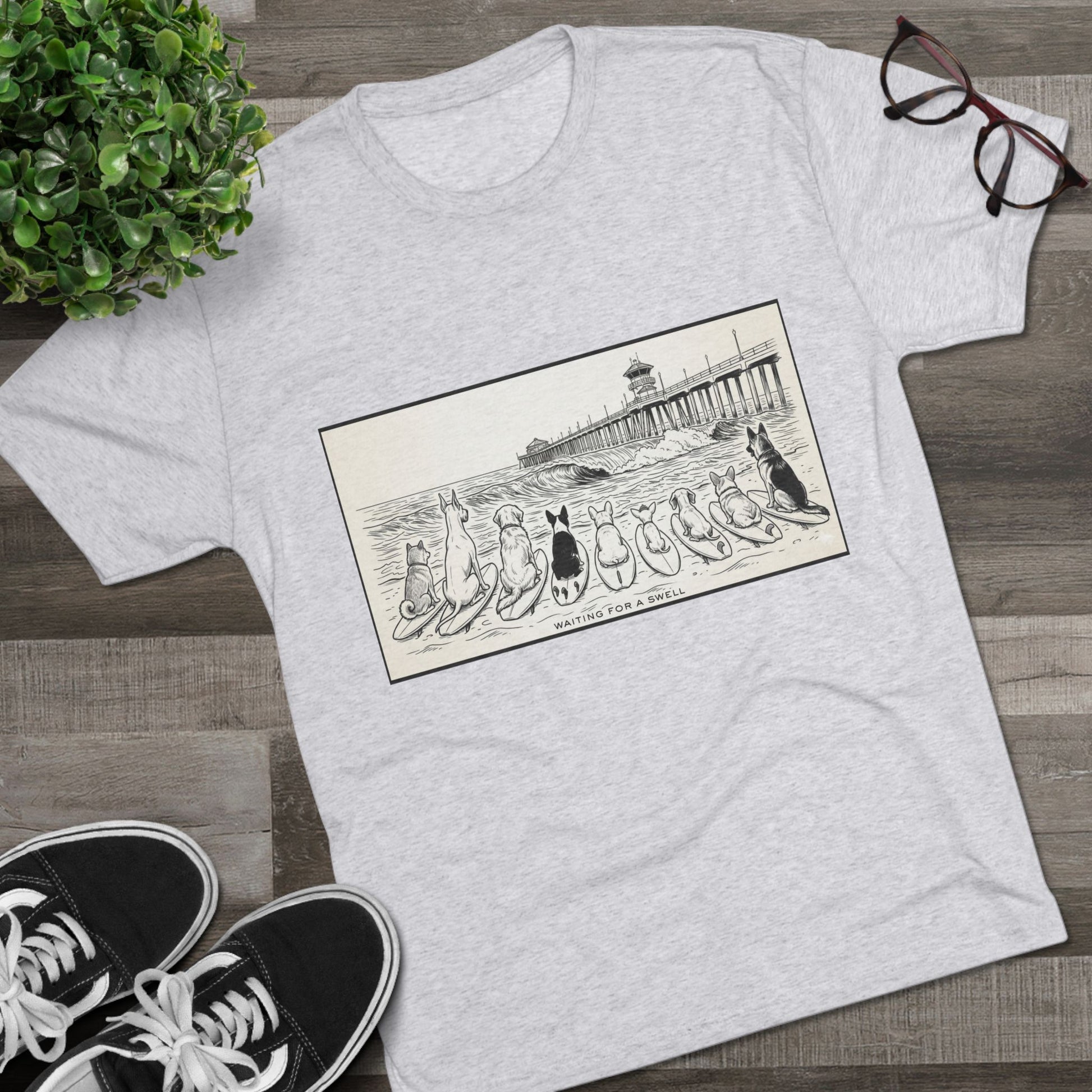 White t-shirt with a pack of dogs on surfboards staring out to sea, with the words Waiting for a Swell' underneaath the dogs and the Huntington Beach pier in the background laying on a wood floor with a pair of Vans, a pair of glasses beside it a plant.