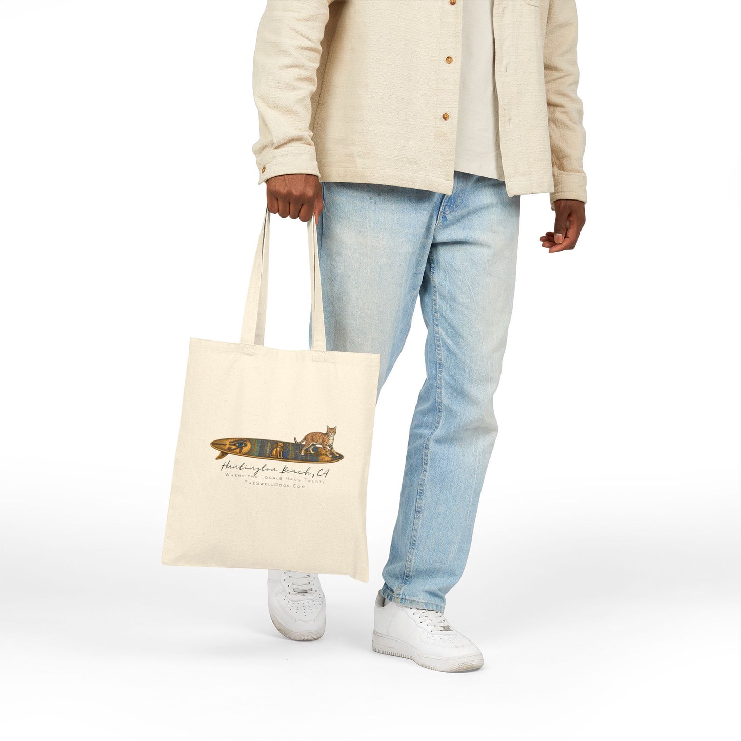 Man carrying a biege tote bag featuring a tabby cat standing on an Egyptian surfboard with "Huntington Beach, CA" text