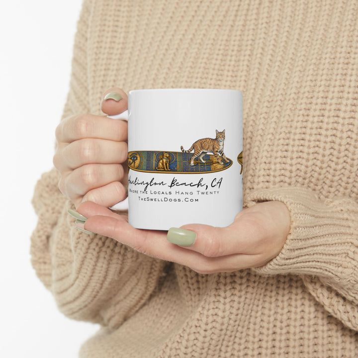 Person holding a white mug featuring a Tabby Cat standing on a Egyptian surfboard with "Huntington Beach, CA" text