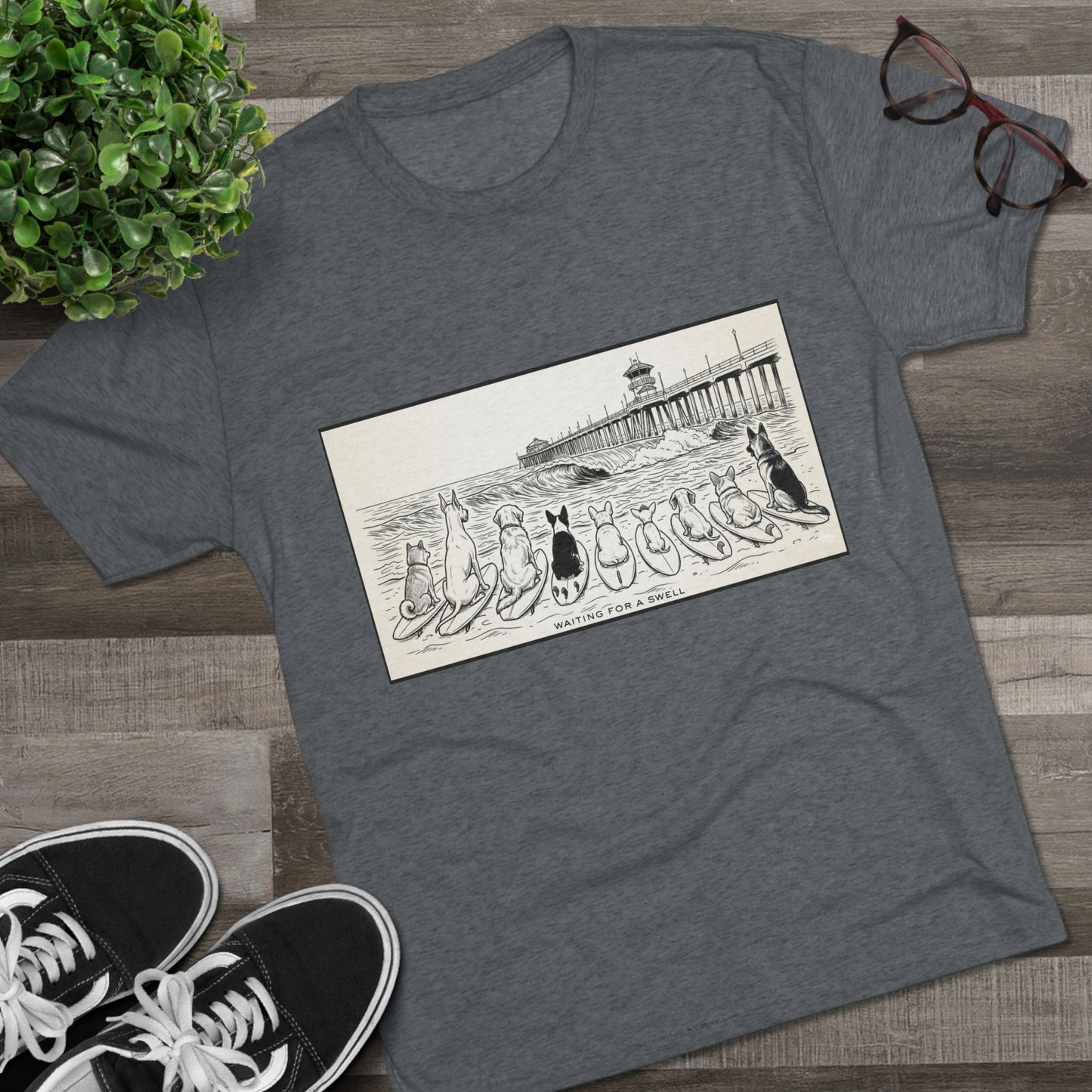 Gray t-shirt with a pack of dogs on surfboards staring out to sea, with the words Waiting for a Swell' underneaath the dogs and the Huntington Beach pier in the background laying on a wood floor with a pair of Vans, a pair of glasses beside it a plant.
