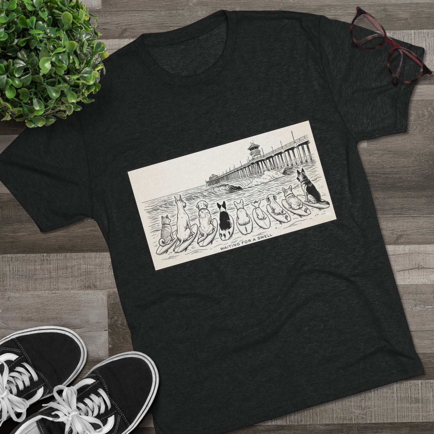 Black t-shirt with a pack of dogs on surfboards staring out to sea, with the words Waiting for a Swell' underneaath the dogs and the Huntington Beach pier in the background laying on a wood floor with a pair of Vans, a pair of glasses beside it a plant.