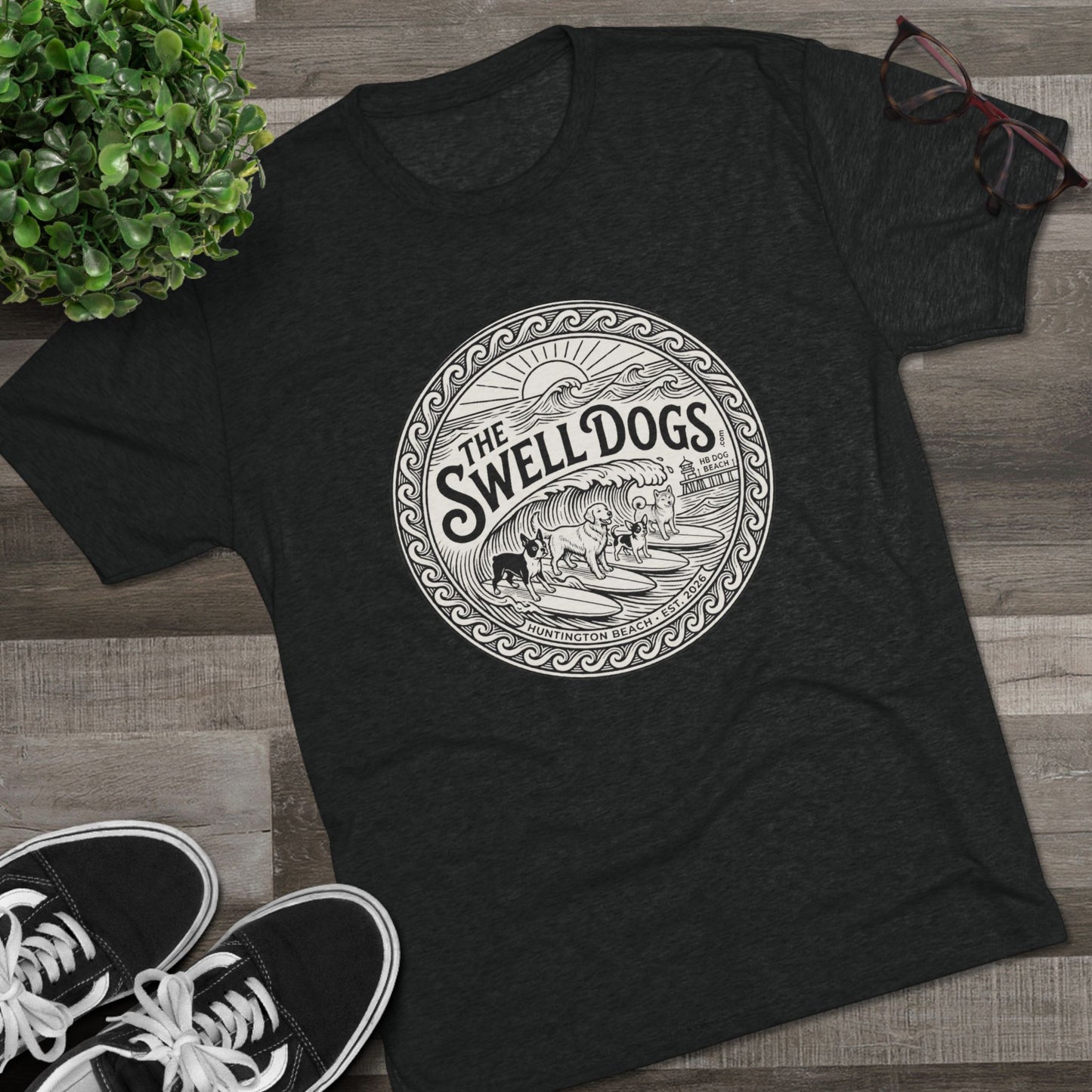 Black  tshirt with The Swell dogs logo on it on a wood floor with a plant and a pair of Vans.
