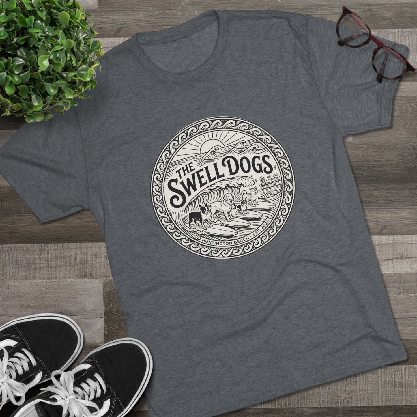 Gray tshirt with The Swell dogs logo on it on a wood floor with a plant and a pair of Vans.