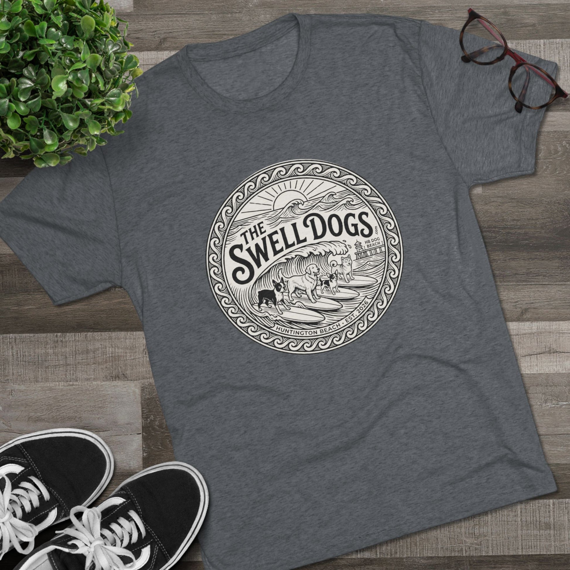 Gray tshirt with The Swell dogs logo on it on a wood floor with a plant and a pair of Vans.