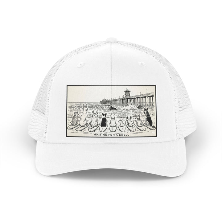 White trucker hat with a picture of a pack of nine dogs sitting on their surfboards, facing the ocean, with the Huntington Beach pier in the background. It says Waiting for a Swell at the bottom.