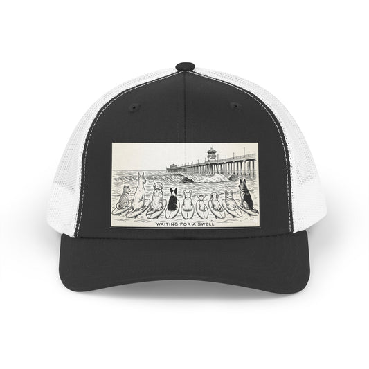 Black and white trucker hat with a picture of a pack of nine dogs sitting on their surfboards, facing the ocean, with the Huntington Beach pier in the background. It says Waiting for a Swell at the bottom.