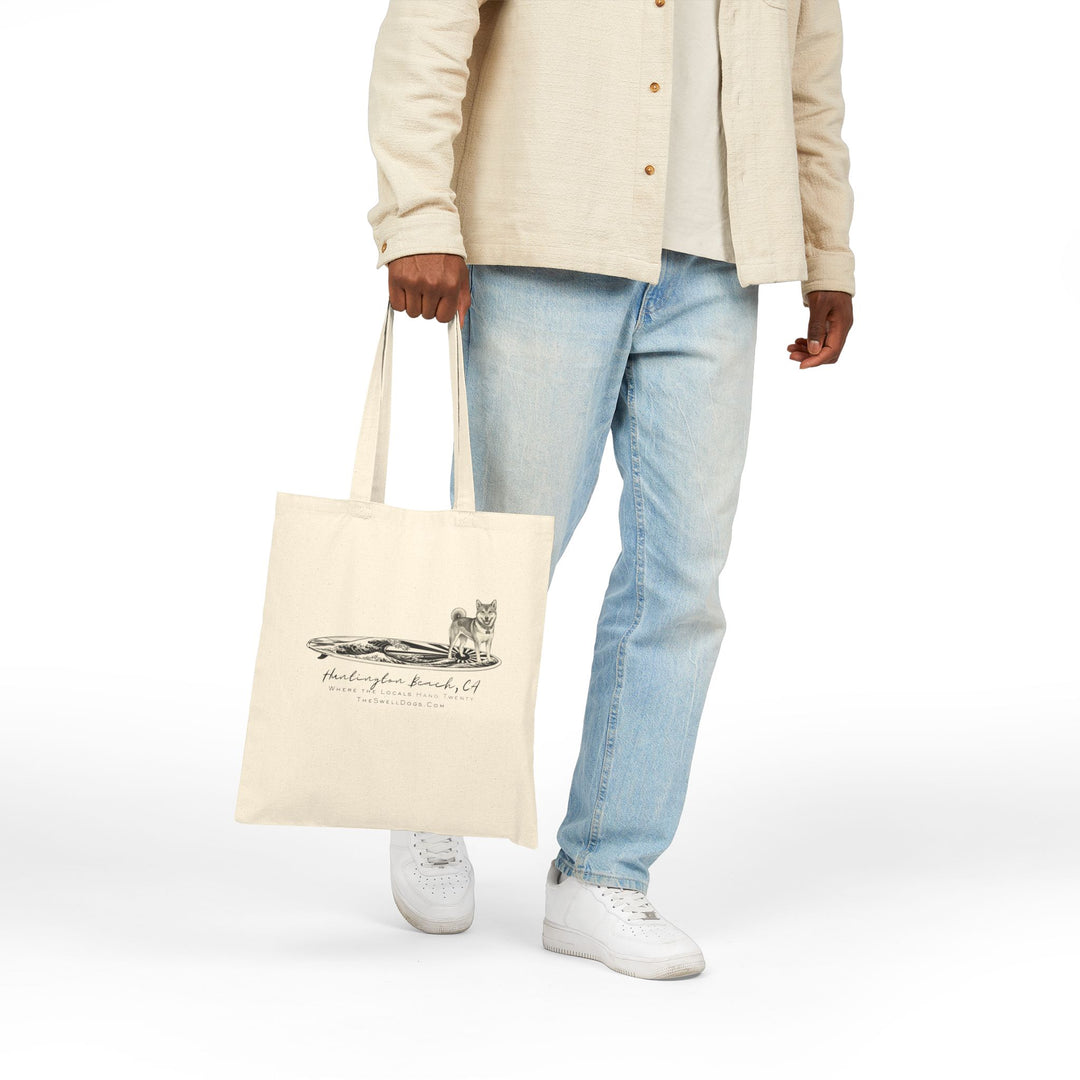 Man carrying a beige tote bag featuring a Shiba Inu dog standing on a Japanese surfboard with "Huntington Beach, CA" text