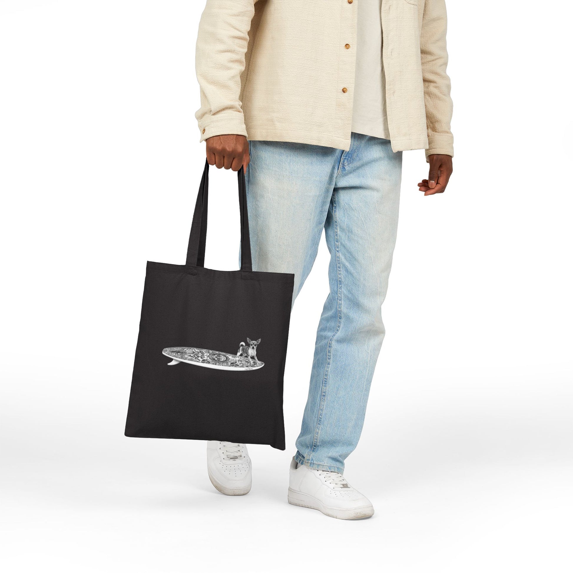 Person holding a blacktote bag with a Chihuahua surfing on a Day of the Dead surfboard.