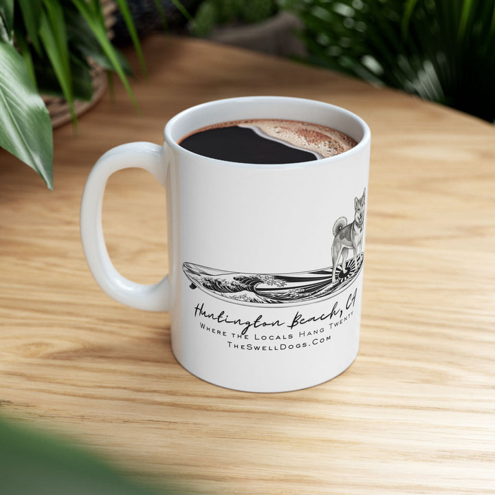 White mug filled with coffee featuring a Shiba Inu standing on a Japanese surfboard with “Huntington Beach, CA” text
