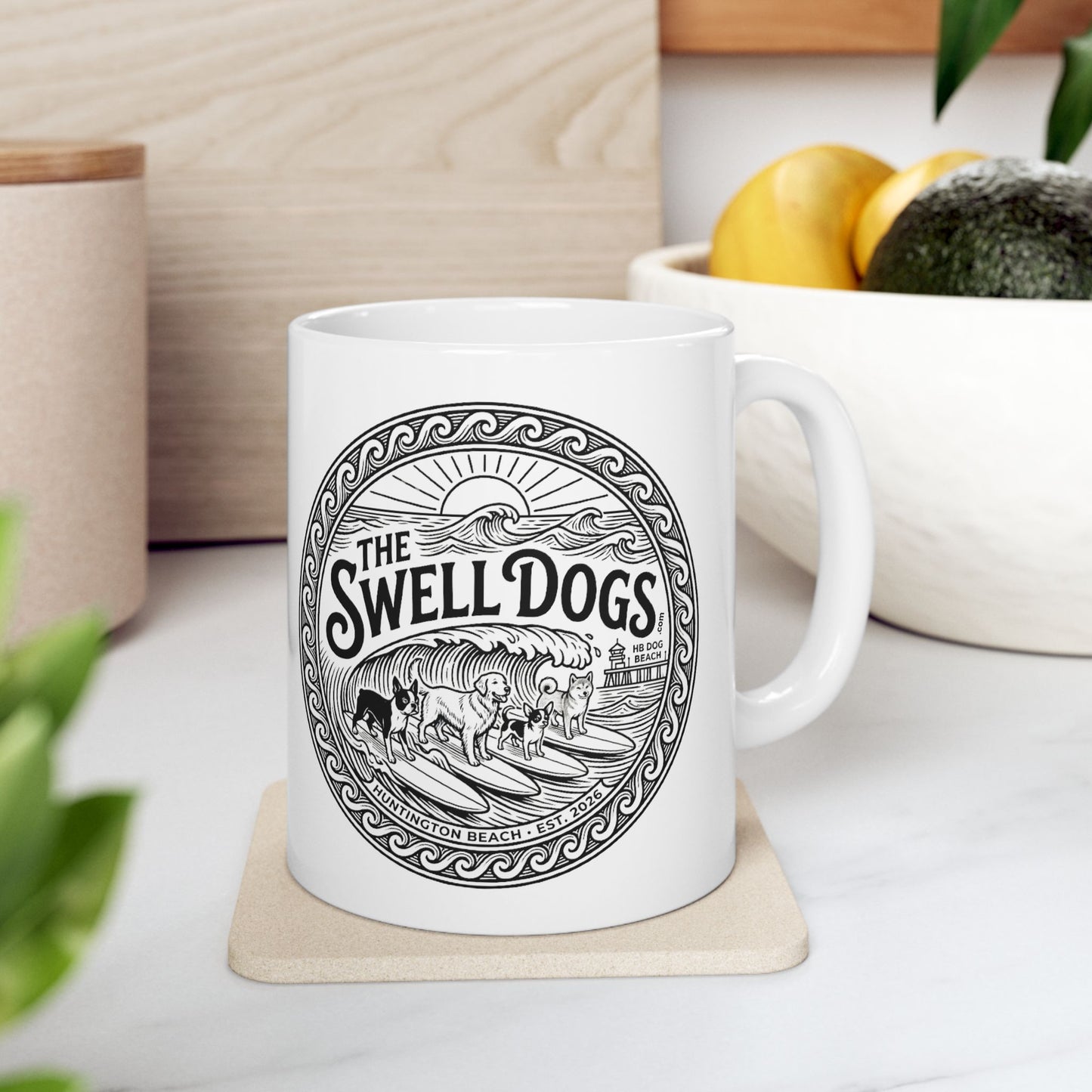 White mug with The Swell Dogs logo on it on a white table with a bowl of fruit next to it.
