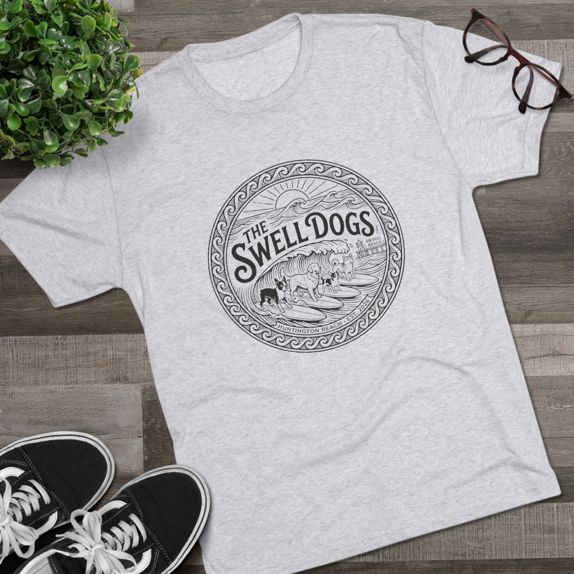 White tshirt with The Swell dogs logo on it on a wood floor with a plant and a pair of Vans.