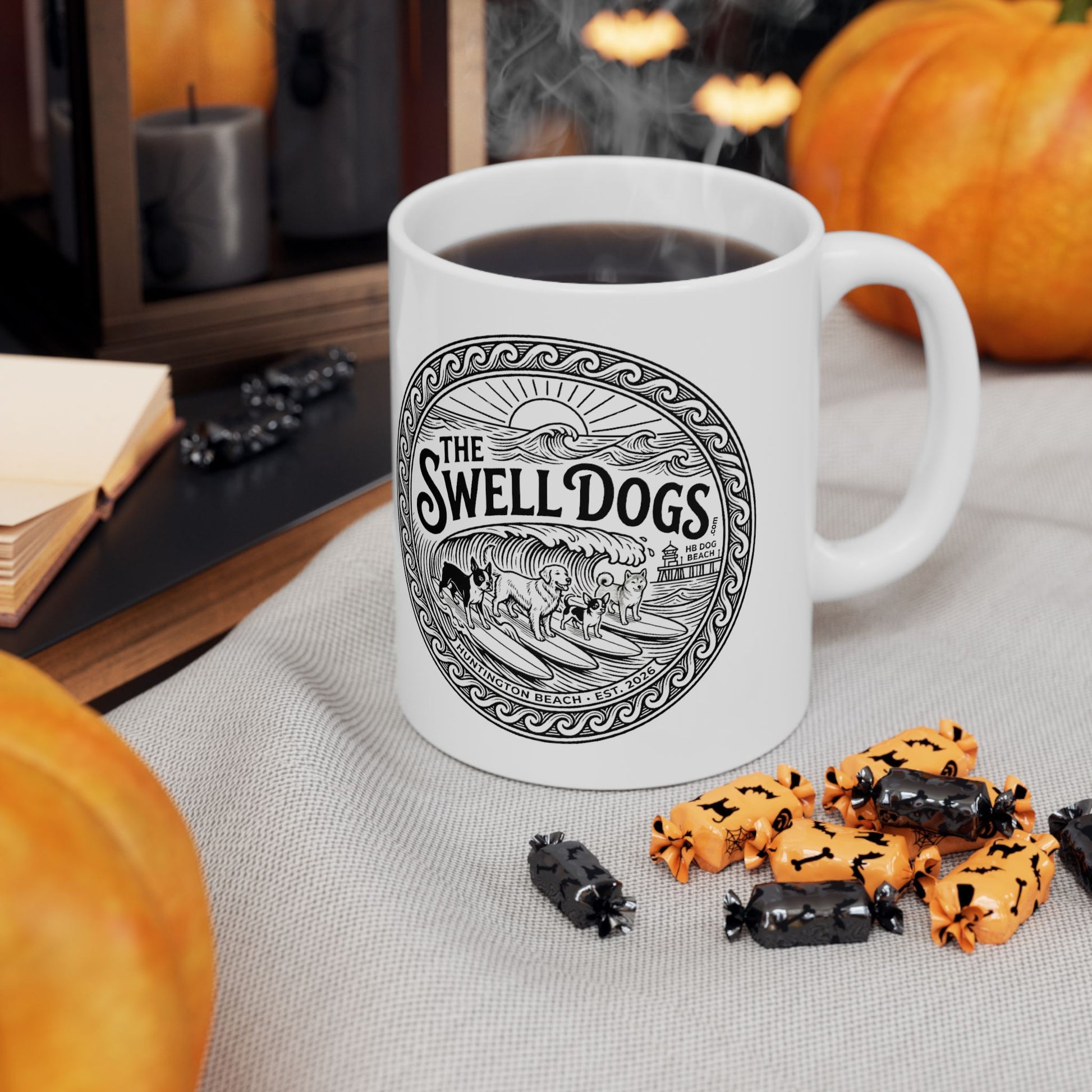 White mug with The Swell Dogs logo on it with pumpkins and halloween candy nearby.
